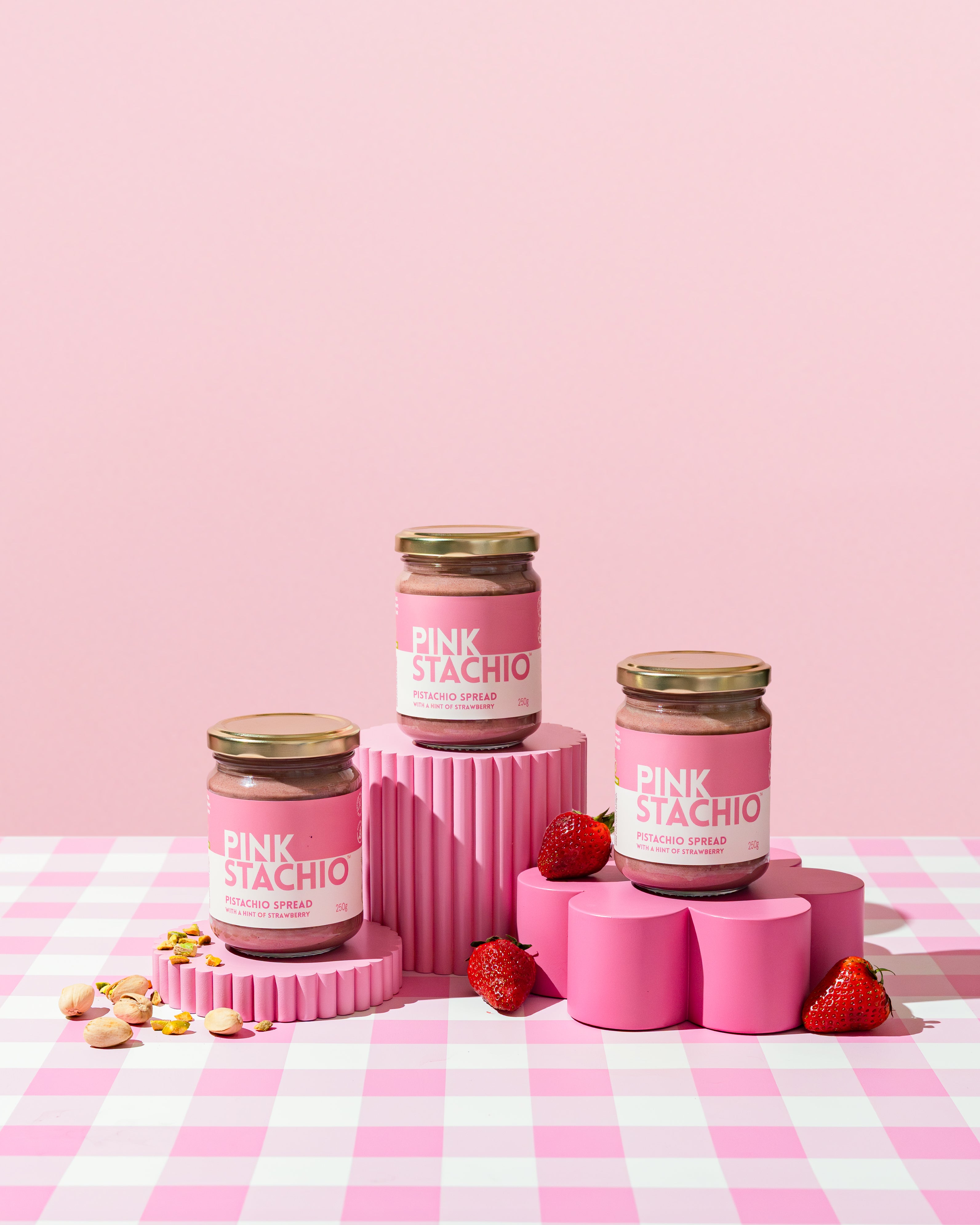 Jars of 'Pink Stachio' on pink stands with strawberries and cookies on a pink and white checkered background.